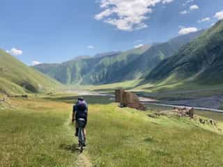 Een fietser rijdt door een groen dal in Georgië, omringd door bergen en ruïnes onder een blauwe lucht.
