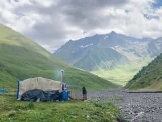 Een geïmproviseerde hut in een groen bergdal in Georgië, met majestueuze bergen op de achtergrond onder bewolkte lucht.