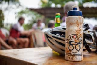 Een modderige drinkenbus met het logo van Veloglobe en op de achtergrond deelnemers die ontspannen op hun fietsreis in Rwanda.